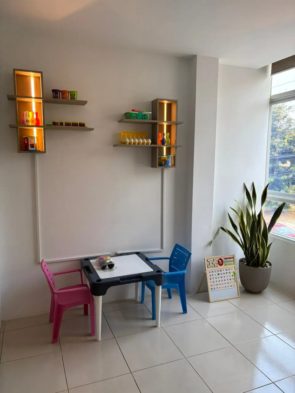 Rincón infantil con mesa de trabajo, sillas de colores y material didáctico en repisas iluminadas.